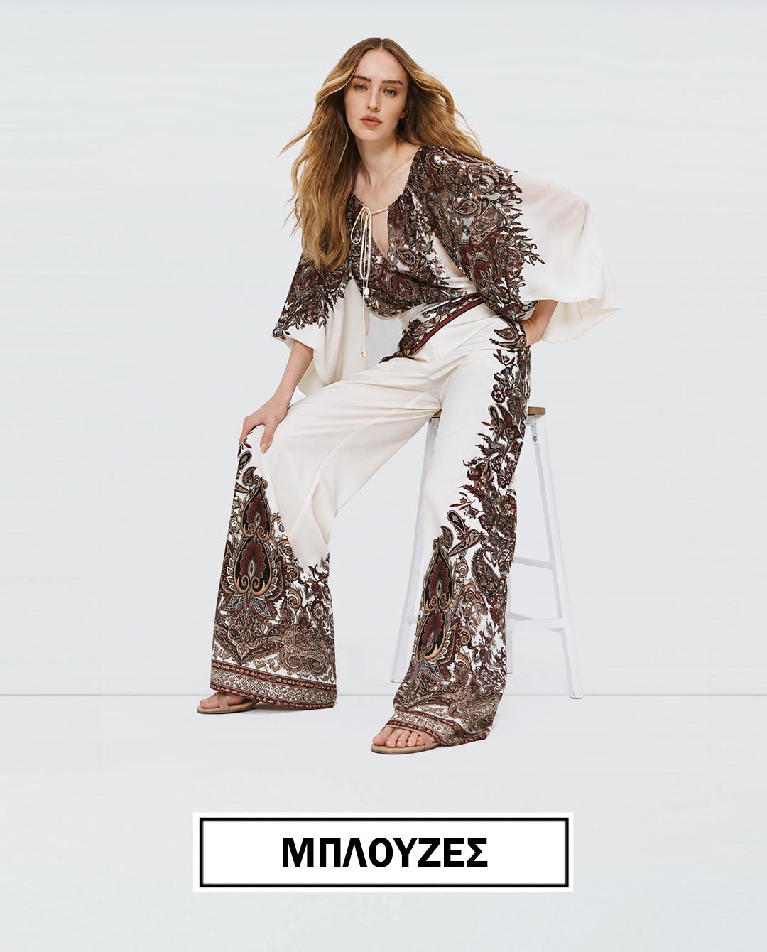 Model wearing a cream bohemian blouse with brown paisley embroidery and matching wide leg pants seated on a stool against a white backdrop as a fashion promo image