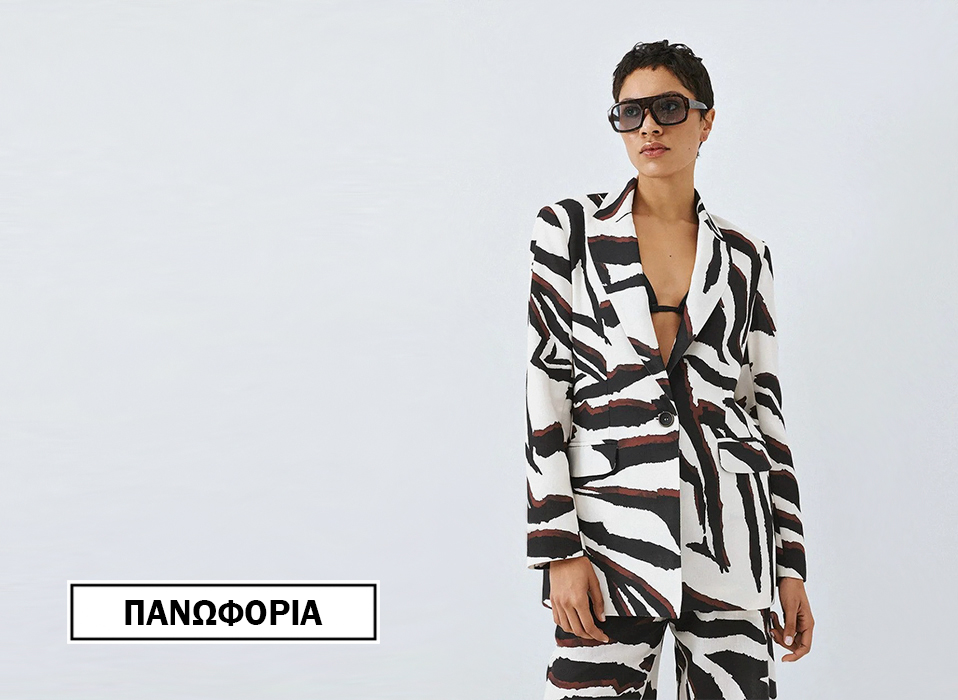 Model wearing a bold black and white zebra print blazer and matching pants with a black top visible at the neckline and dark sunglasses standing against a light background bottom left logo in a bordered box displays Greek text